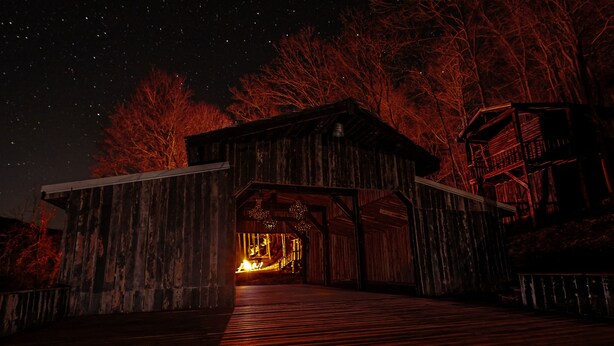 A rustic wooden structure with a wooden deck leading to a warmly lit interior, surrounded by trees. - Glamping Cabin | Huge Window | Tree House Feel (Green Township)