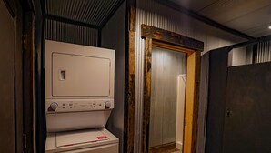 A compact laundry room with a front-loading washer and dryer, wooden flooring, and a door leading to another room. - Glamping Cabin | Huge Window | Tree House Feel (Green Township)