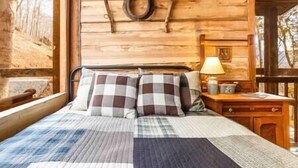 A wooden cabin room with a bed, checkered bedding, a nightstand with a lamp, and a window with a view of trees. - Glamping Cabin | Huge Window | Tree House Feel (Green Township)