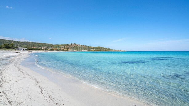 Ein Strand mit klarem türkisblauem Wasser, einem Sandstrand und einer Hügelkuppe mit Gebäuden im Hintergrund.
