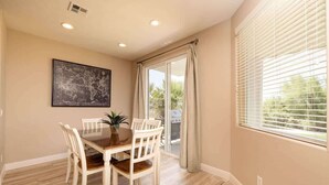 A dining area with a wooden table and chairs, a potted plant, a framed map on the wall, and a view through a window with blinds.