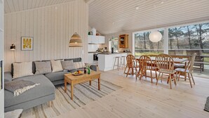 Een ruime woonkamer met een bank, salontafel en houten stoelen. Een keuken met witte kasten en een groot raam is zichtbaar.