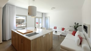 Une cuisine moderne dotée d’un îlot central, de plans de travail blancs et d’armoires en bois. On y trouve un coin repas avec une table et des chaises blanches, ainsi qu’un salon avec un canapé blanc et des coussins rouges.