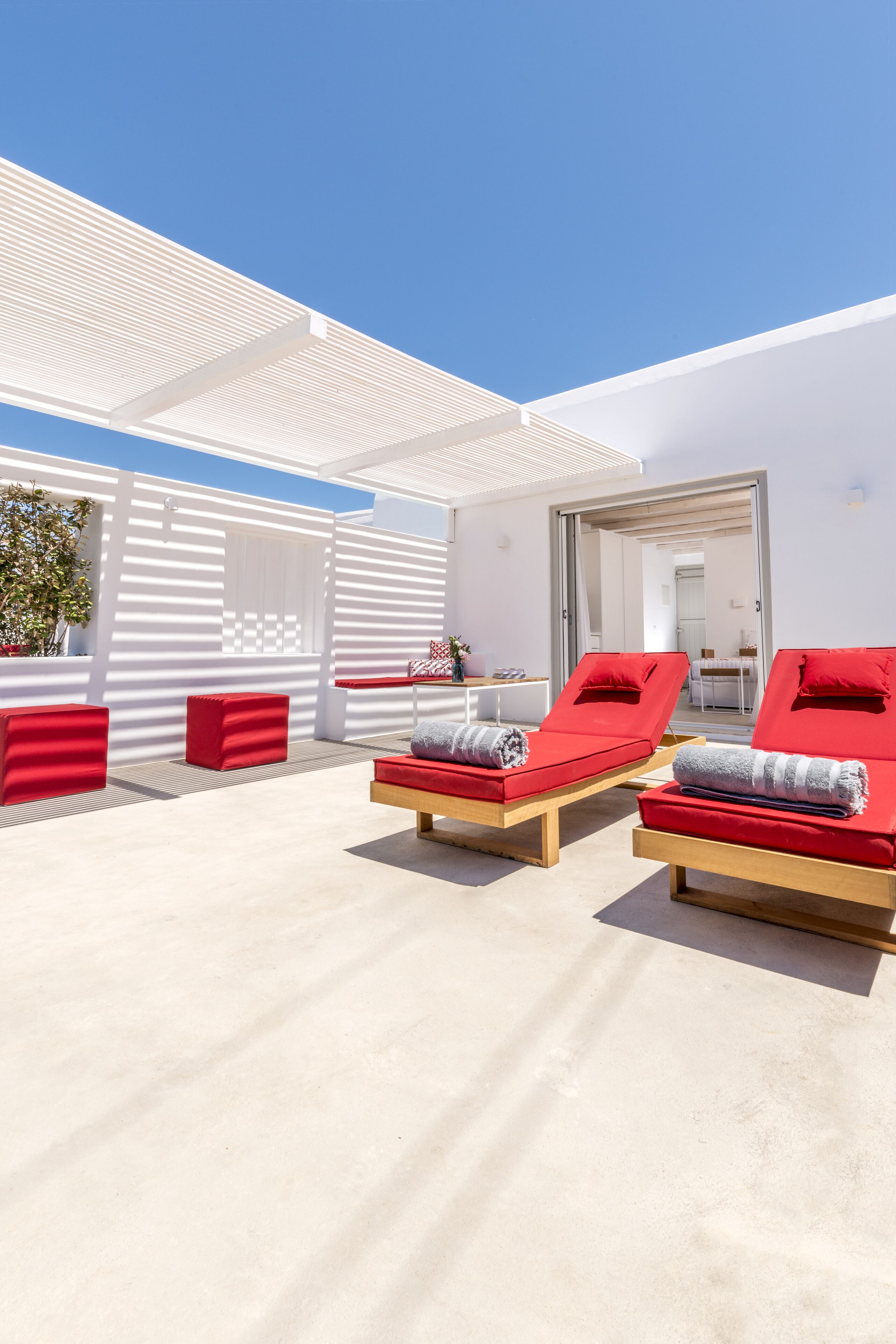 A sunlit outdoor lounge area with two red lounge chairs, a wooden bench, and a white wall with a window.