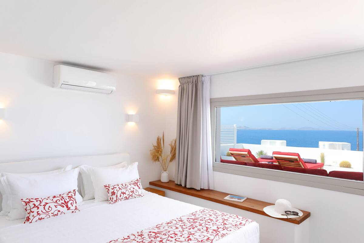 Une chambre d’hôtel avec un lit, une fenêtre donnant sur la mer, une table de chevet avec un chapeau, et un climatiseur fixé au mur.