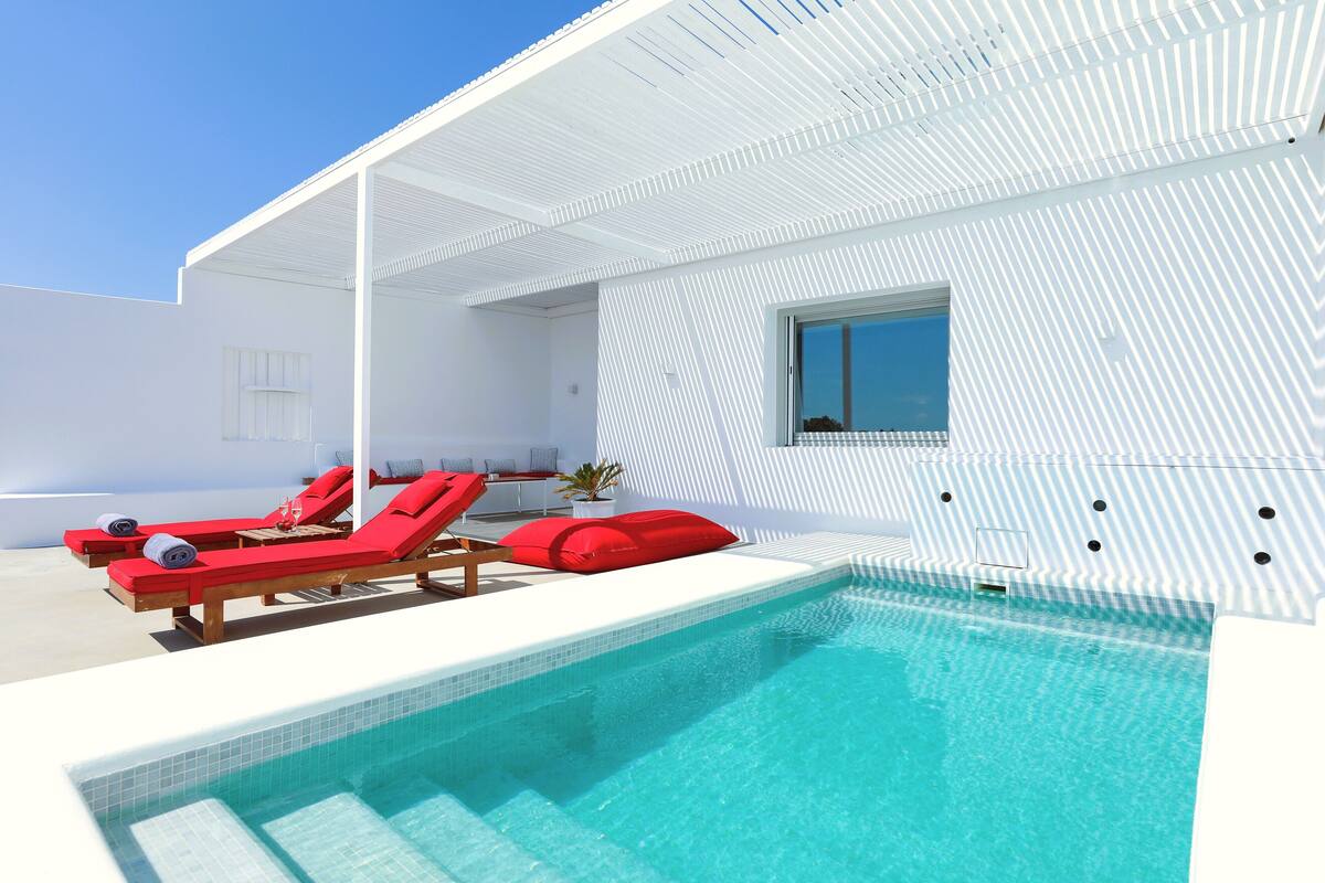 Un espace piscine moderne avec deux chaises longues rouges et une petite table, une piscine à l’eau d’un bleu limpide et une façade blanche ornée d’un motif rayé original.