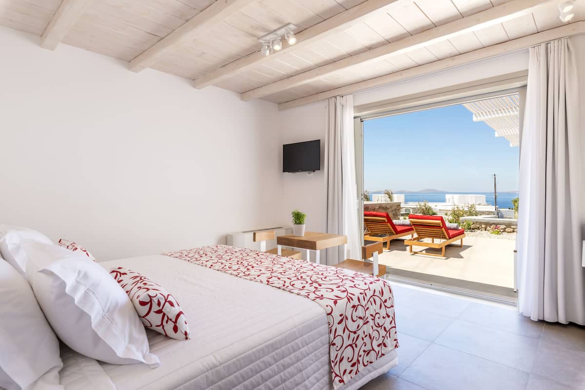 Une chambre avec un lit, une télévision, un balcon avec des chaises et une vue sur la mer.