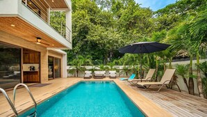 A modern outdoor swimming pool with wooden decking, lounge chairs, and a small kitchen area. - 3 King Suites | Pool, Outdoor Oasis | Casa Coralis (Potrero)