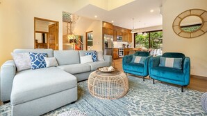 A spacious living room with a grey sectional sofa, two teal armchairs, and a round coffee table. A kitchen is visible in the background with wooden cabinets and a large window. - 3 King Suites | Pool, Outdoor Oasis | Casa Coralis (Potrero)