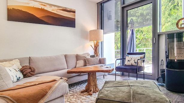 Uma sala de estar moderna com um sofá, uma mesa de centro e uma poltrona. Há uma janela grande com vista para o verde lá fora.