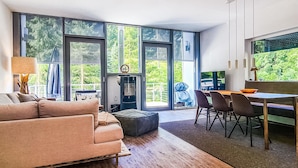 Uma sala de estar moderna com um sofá, uma mesa de jantar com cadeiras e grandes janelas com vista para o verde.