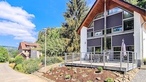 Um edifício moderno de dois andares com varanda, rodeado por vegetação e um céu limpo.