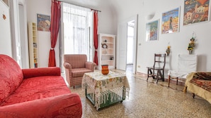 Ein Wohnzimmer mit einem roten Sofa, einem karierten Sessel, einem kleinen Tisch mit einer Vase und einem weißen Stuhl. Es gibt Bücherregale und eine Tür zu einem anderen Raum.