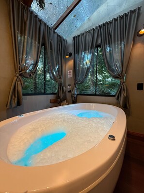 A large bathtub with bubbles, a window with curtains, and a ceiling light.