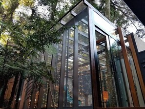 A modern glass structure with vertical supports, surrounded by dense foliage.