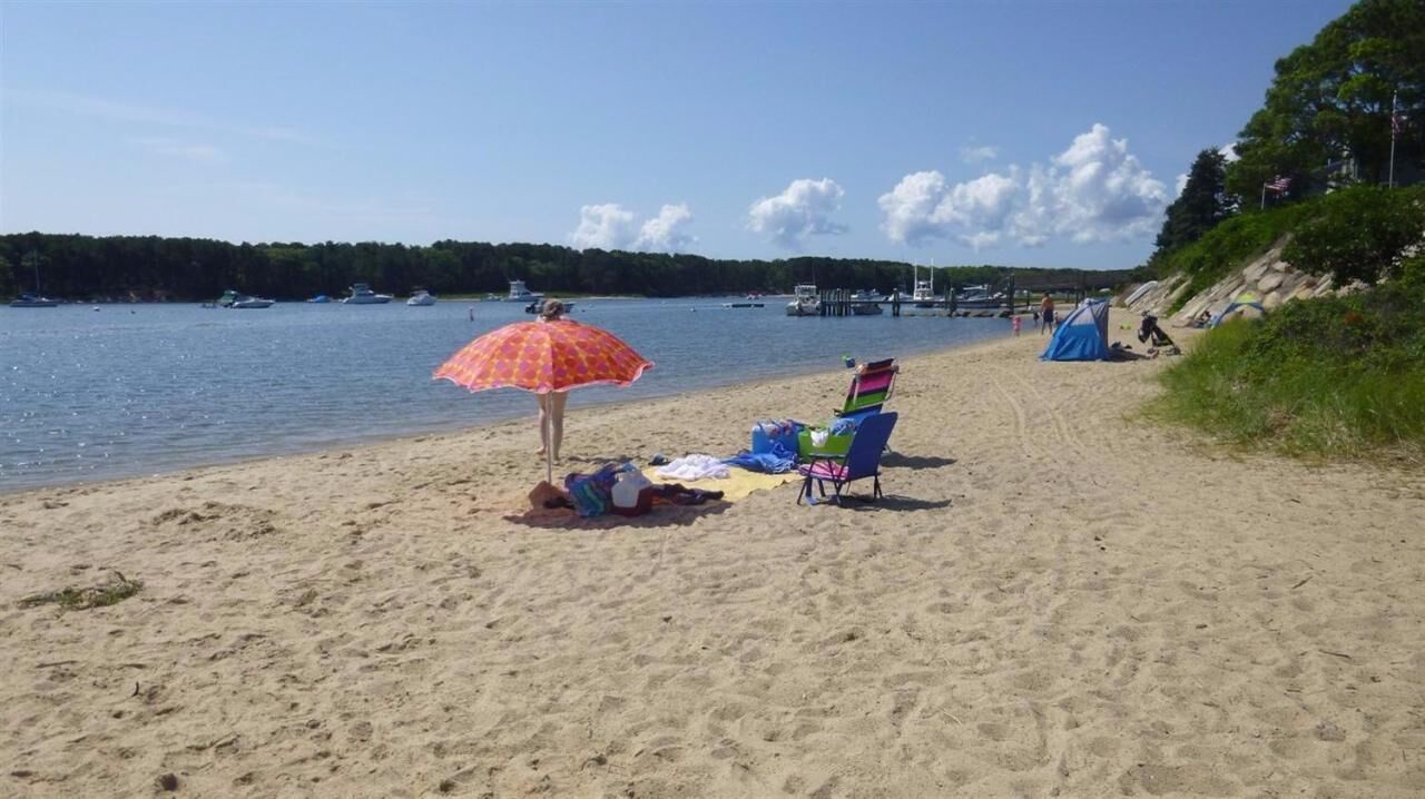 Ein Strand mit sandigem Ufer, eine Frau unter einem Sonnenschirm und ein Zelt im Hintergrund.