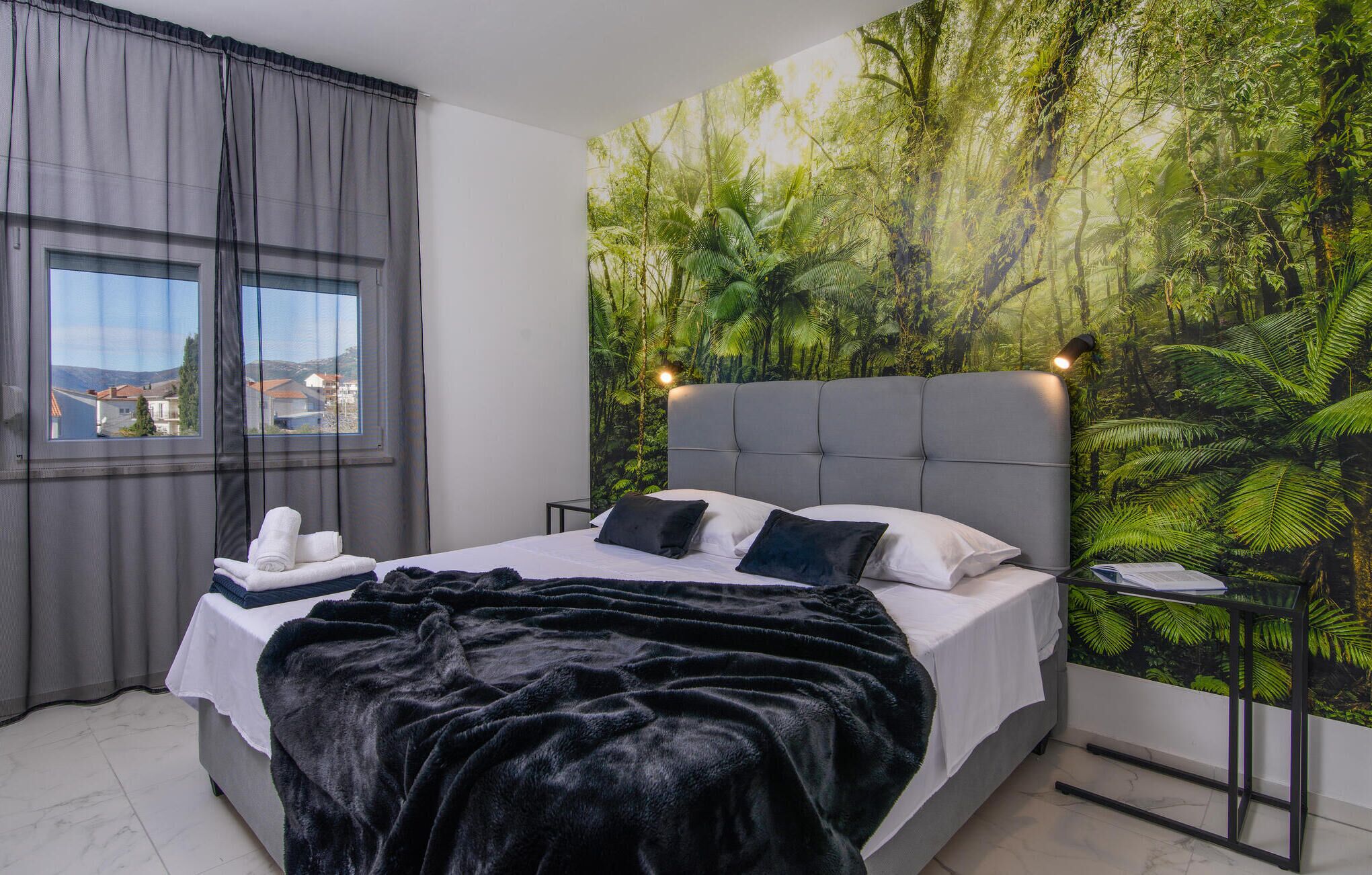A bedroom with a large bed, grey curtains, a view through the window, and a tropical forest mural on the wall.