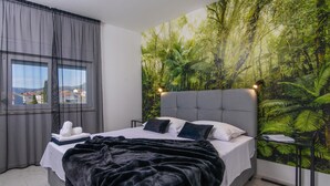 Quarto com cama grande, cortinas cinzas, vista pela janela e um mural de floresta tropical na parede.