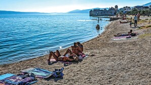 Uma praia com pessoas tomando sol e nadando, um edifício histórico ao fundo, e um céu azul límpido.