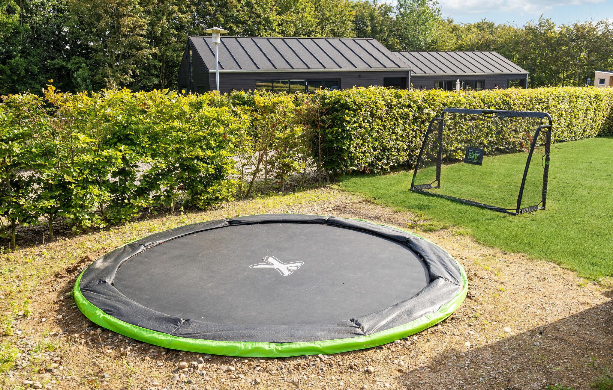 A trampoline in a yard with a hedge, a swing set, and a building in the background.