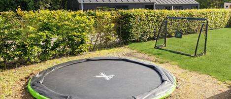 A trampoline in a yard with a hedge, a swing set, and a building in the background.