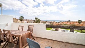 Eine Terrasse mit Holztisch und -stühlen, einer weißen Wand und Blick auf eine Küstenstadt.