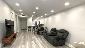 A spacious living area with a grey sectional sofa, a flat-screen TV, and a wooden entertainment unit. - FL Beach Get-Away! 1BD/1BA in 2BD/2BA Available in South Florida (Delray Beach)