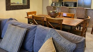 Uma sala de estar com sofá, almofadas estampadas, uma mesa de jantar, cadeiras e uma cozinha ao fundo, com armários e eletrodomésticos.