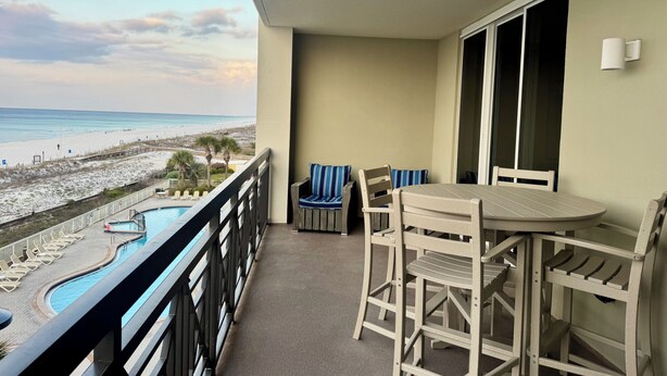 Uma varanda com vista para a praia, piscina e área de refeições com cadeiras e mesa.
