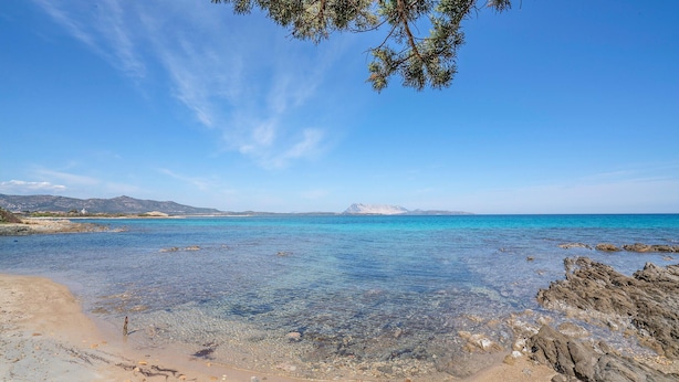 Une plage côtière aux eaux claires, parsemée de rochers et avec une montagne qui se profile à l’horizon.