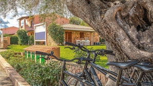 Un vélo est garé dans un jardin luxuriant, avec un mur de pierre et un grand tronc d’arbre au premier plan.