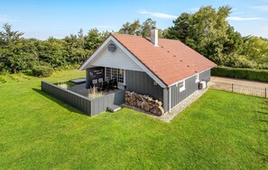 Une maison moderne avec un toit en pente, une terrasse dotée de mobilier d’extérieur et une pile de bois de chauffage.