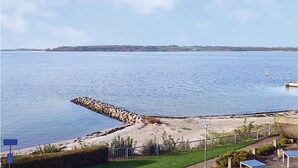 Vista costanera amb un pont de pedra que s'enduu cap a aigües tranquil·les, una zona verda amb una tanca i una riba llunyana.
