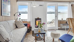 Ein gemütliches Wohnzimmer mit einer Couch, einem kleinen Tisch mit Blumen in einer Vase, einem Kamin und Blick ins Freie durch große Schiebetüren.