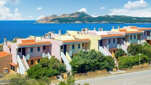 Une zone résidentielle côtière, avec des maisons aux couleurs pastel, une mer d’un bleu limpide et un paysage montagneux en arrière-plan.