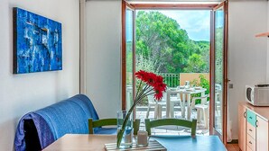 Une pièce comprenant une table à manger, des chaises, un canapé, un tableau et une vue sur la verdure, visible à travers une porte vitrée.
