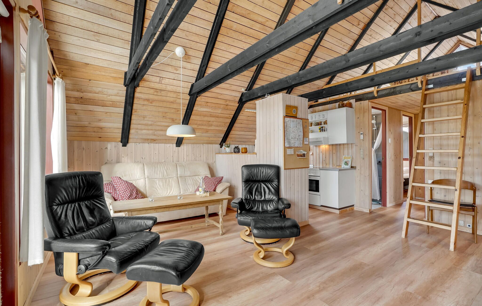Ein gemütlicher Raum mit Holzbalkendecke, einer Couch, zwei schwarzen Ledersesseln, einem kleinen Tisch und einer Hockerleiter.