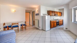 Cocina compacta con refrigerador, estufa y gabinetes de madera. Cuenta con una zona de comedor con mesa y sillas, y un sofá.