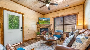 A cozy cabin interior with a stone fireplace, a ceiling fan, and a large window with blinds. - ★Modern★ Family Friendly Cabin in Pigeon Forge - Hot Tub Game Room Fire Pit (Pigeon Forge)