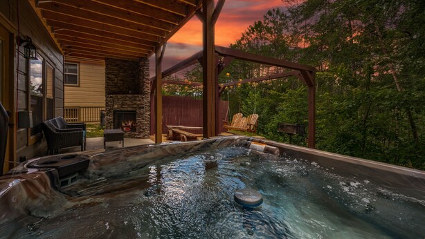 A hot tub under a wooden pergola with a stone fireplace, surrounded by outdoor furniture and a stone wall. - ★Modern★ Family Friendly Cabin in Pigeon Forge - Hot Tub Game Room Fire Pit (Pigeon Forge)