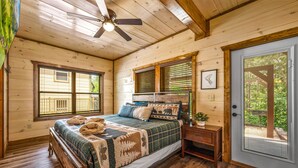A wooden cabin bedroom with a bed, a nightstand, a ceiling fan, and a door leading to a deck. - ★Modern★ Family Friendly Cabin in Pigeon Forge - Hot Tub Game Room Fire Pit (Pigeon Forge)