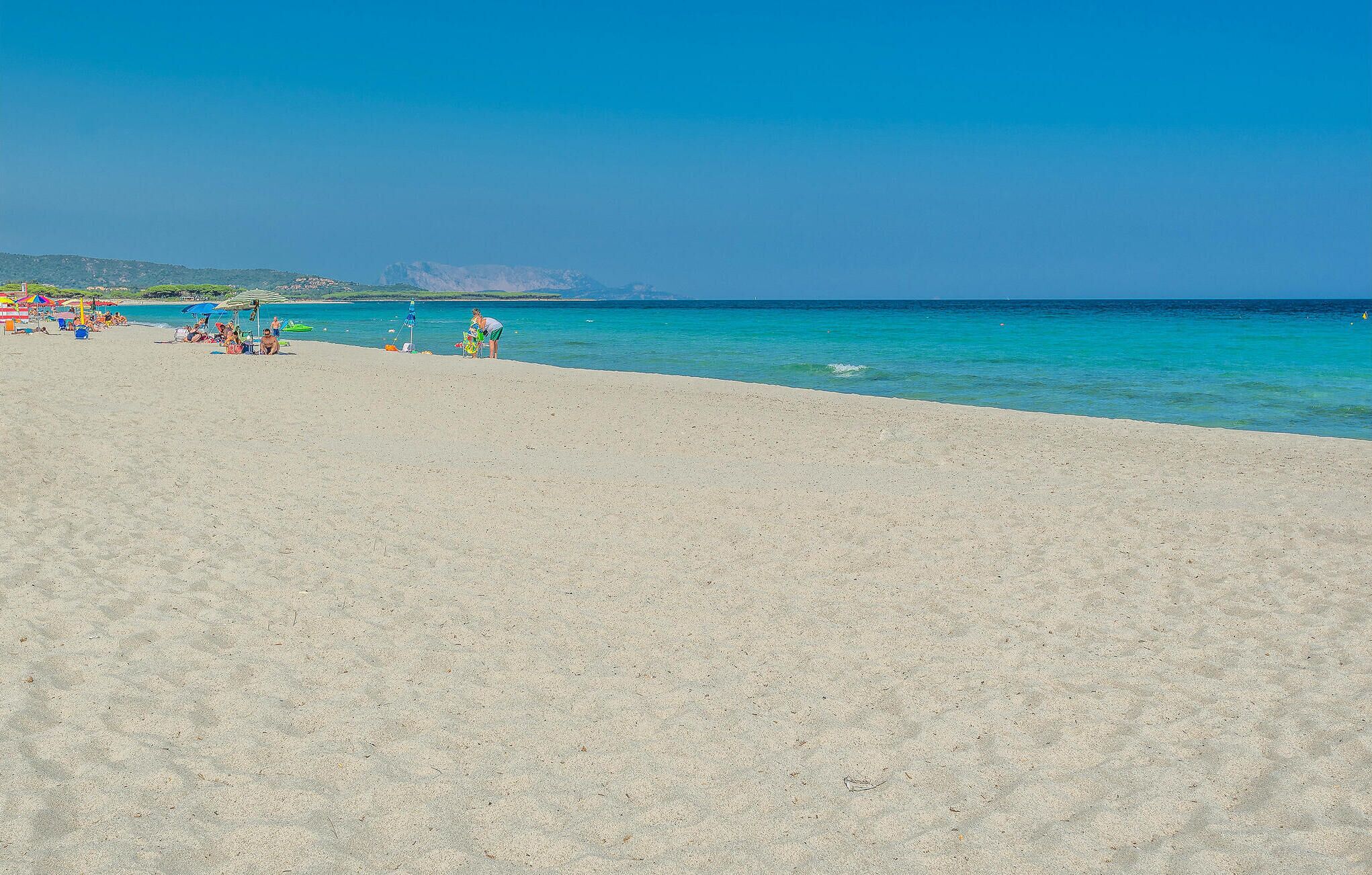 Ein Strand mit klarem, türkisblauem Wasser und einem sandigen Ufer. Liegestühle und entspannte Menschen sind zu sehen.