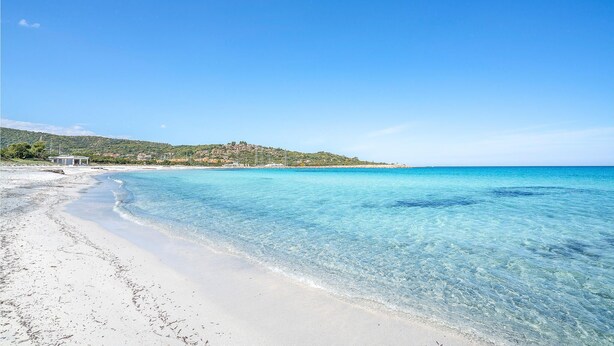 Ein Strand mit klarem türkisblauem Wasser, einem Sandufer und einem Hügel mit Gebäuden im Hintergrund.