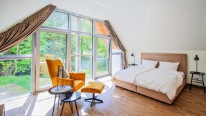 Un dormitorio con un ventanal grande, una cama, un sillón, una mesita pequeña y una lámpara.