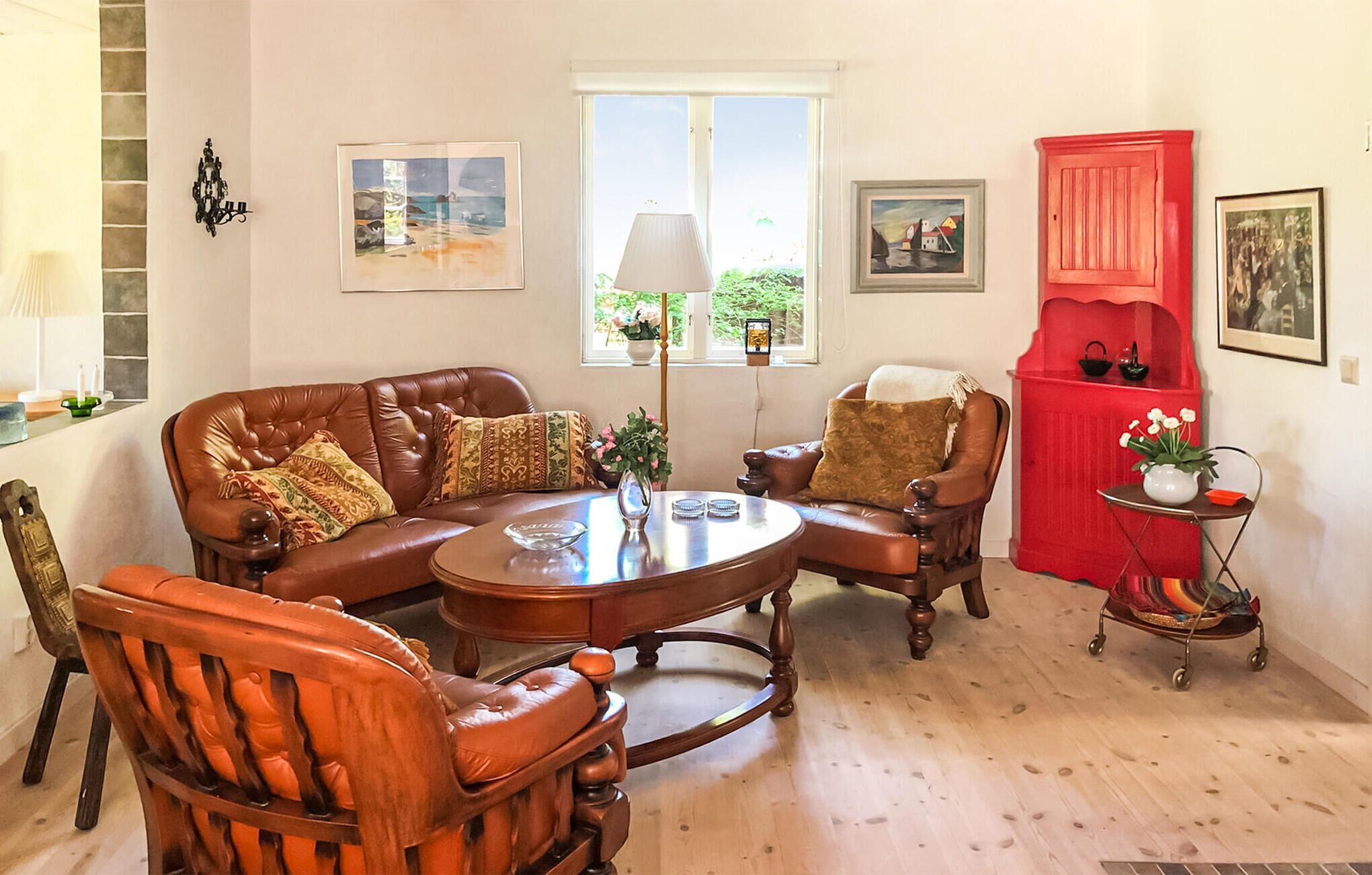 A living room with a brown leather sofa, two armchairs, a wooden coffee table, and a red cabinet. There are framed pictures on the walls and a window with a view of greenery outside.