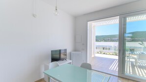 Une pièce moderne et minimaliste, dotée d’une table en verre, de chaises blanches, d’un téléviseur à écran plat et d’une grande porte-fenêtre coulissante donnant sur un balcon extérieur avec vue sur une colline et des bâtiments.