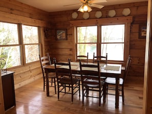 A wooden cabin interior with a dining table and chairs, large windows, and a ceiling fan.