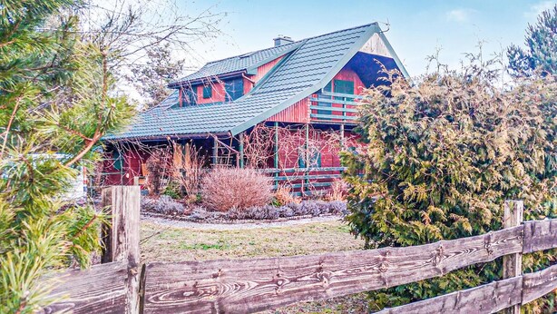 Une grande maison de style rustique, dotée d’un toit à forte pente, entourée d’une clôture en bois et de divers arbustes.