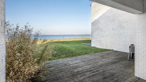 Blick von einer überdachten Terrasse mit Holzdeck auf eine Rasenfläche und einen Strand.