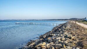 Eine Küstenansicht mit felsiger Küstenlinie, einem sich ins Wasser erstreckenden Steg und einer in der Ferne liegenden Stadtlandschaft.